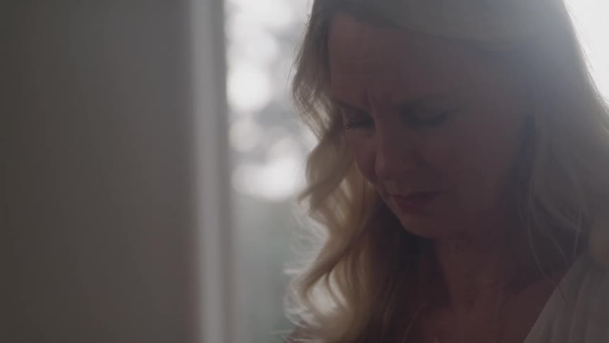 Woman with a sad expression looks down under soft light near the window