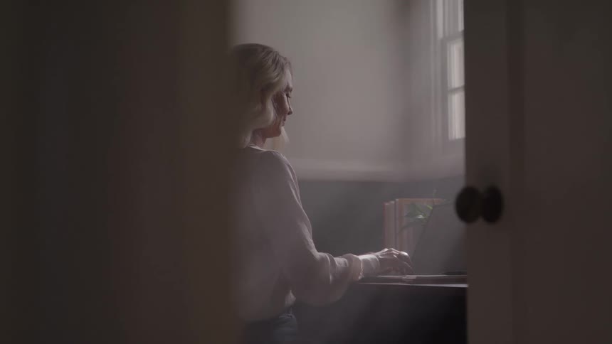 Woman types on laptop at desk with books and plant in naturally lit setting