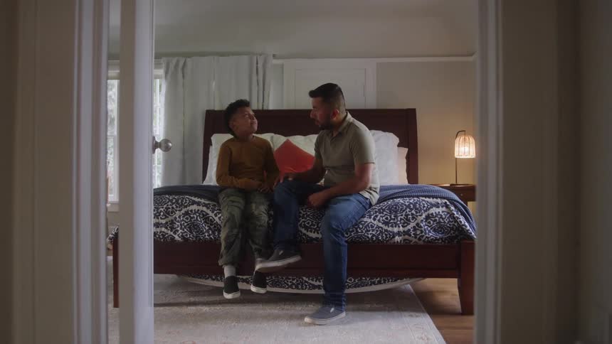 A man in a green shirt and jeans sits on a bed next to a young boy in a brown shirt and khaki pants engaging in a conversation in a bright bedroom seen from an open doorway