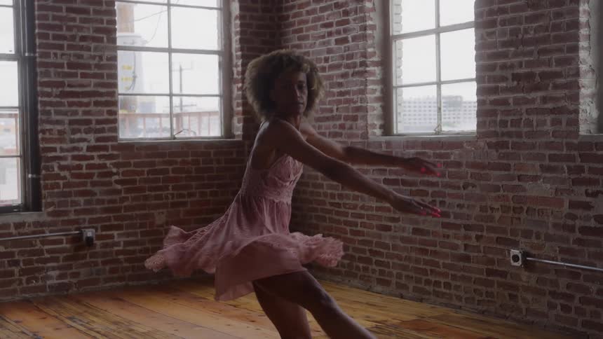 Woman dances with raised leg and extended arms in room with large windows and brick wall