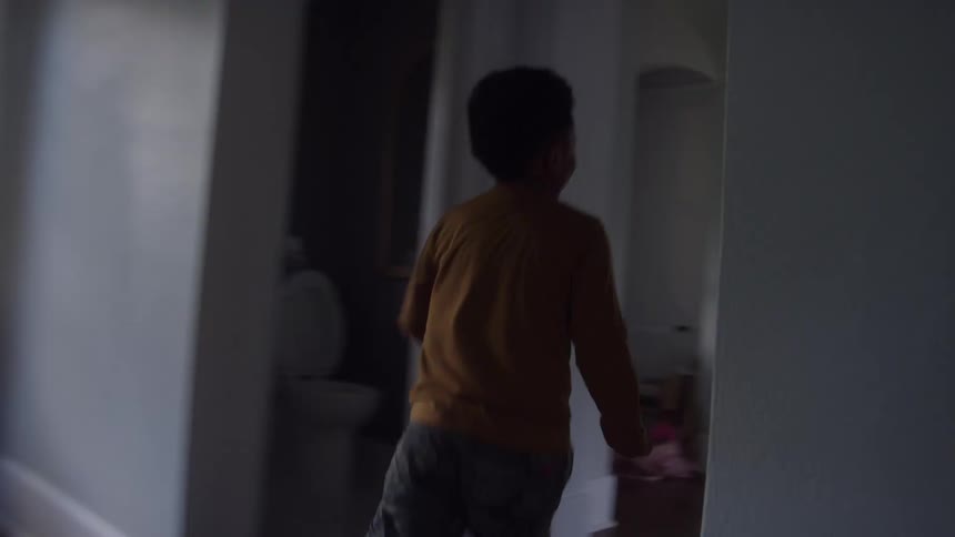 Black young boy running over many rooms of a house and then jumping on the arms of a white man sitting next to a black woman and other people lying on a couch indoors during day time