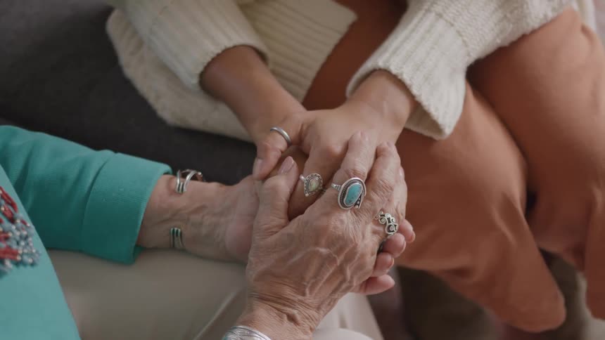 A close view shows the hands of an older woman wearing silver jewelry and a blue top gently being held by the hands of a younger woman wearing a cream sweater and light brown pants
