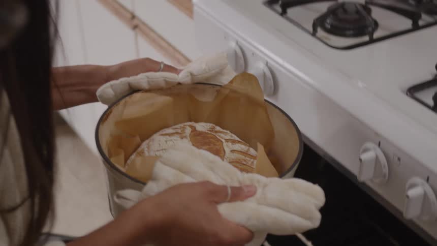 Goldencrusted bread is removed from gas oven with oven mitts in pot lined with parchment paper