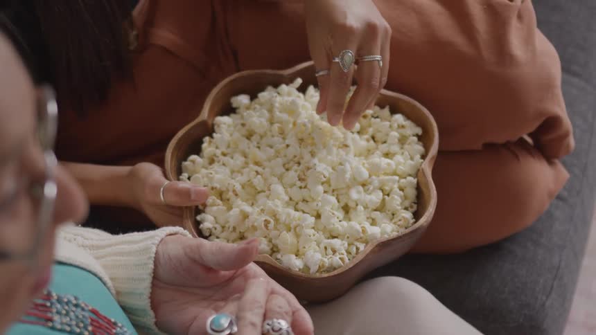 Two women sit comfortably on a couch in the living room sharing a bowl of popcorn as they watch a movie together the midday light filtering softly through the windows