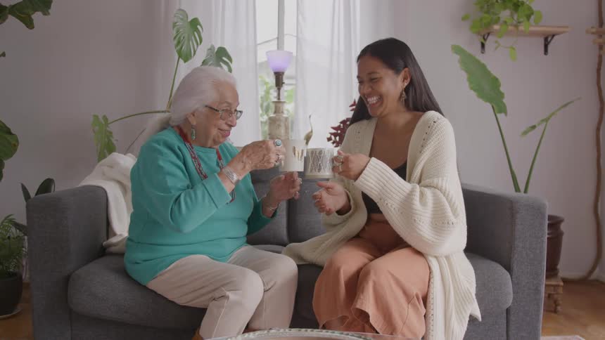 A young woman and an elderly woman laugh joyfully while sitting together on a couch holding mugs in a bright plant filled room