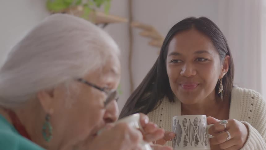 Young woman and elderly lady drinking a beverage from a cup and smiling indoors during day time