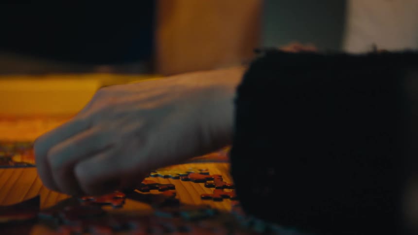 Hand fits puzzle piece on wooden table with yellow box in background and blurred figure beside