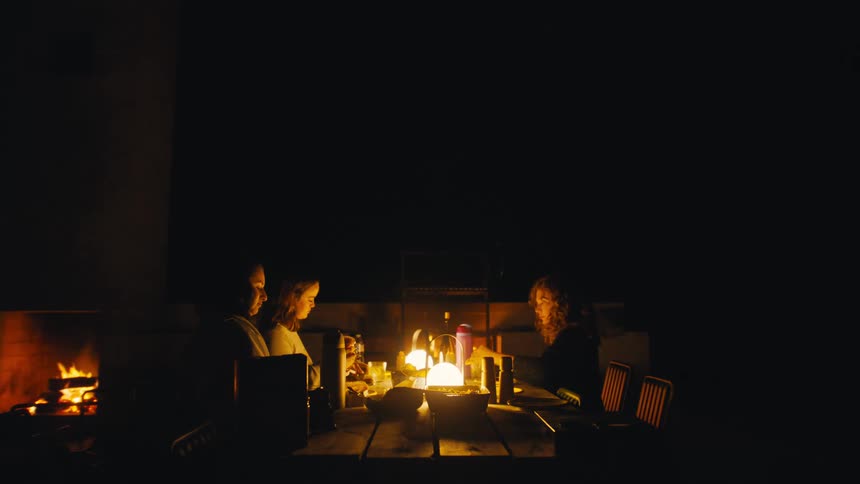 People share a meal by lantern and fire at a wooden table