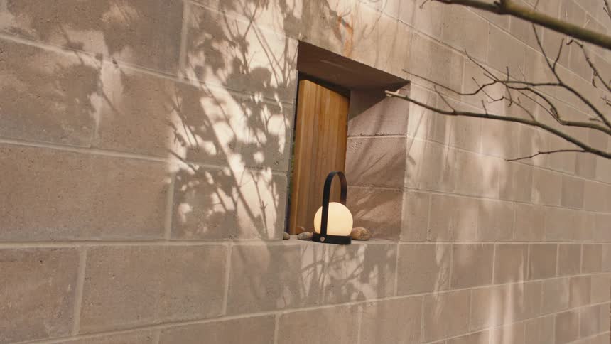 A glowing white orb lamp with a black handle sits in a small square window niche on a concrete block wall as a woman briefly appears and walks through a wooden door