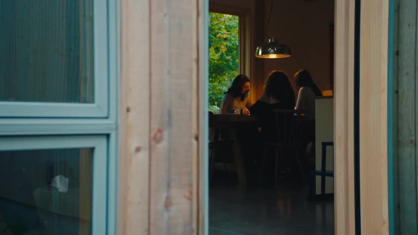 People gather around table lit by hanging lamp in room with window facing green area
