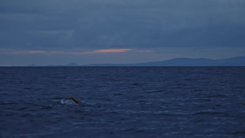 A person wearing a white and orange swim cap swims the freestyle stroke in the choppy dark blue ocean with hazy mountains visible under a dramatic cloudy sky at dawn or dusk