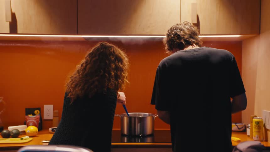From behind a young couple cooks together in a modern warmly lit kitchen with the woman stirring a steaming pot on the stove