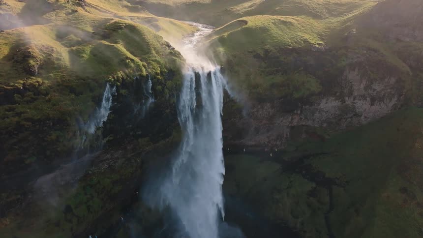 An aerial view shows a massive powerful waterfall plummeting down a sheer cliff face surrounded by vibrant green moss and hills with mist rising from the bottom in the bright sunshine