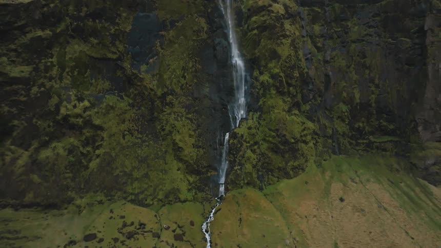 A high thin waterfall cascades down a rugged moss covered cliff face flowing into a rocky stream bed in the grassy foreground under a cloudy sky