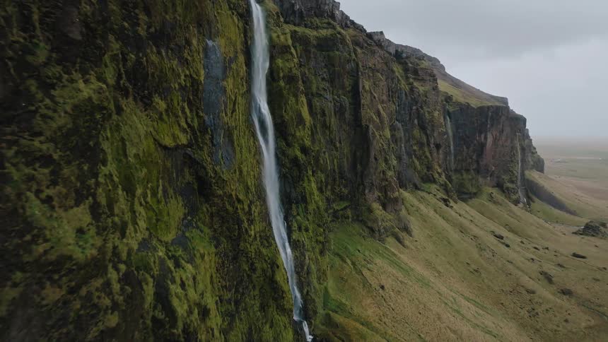 A high angle aerial view moves past a thin tall waterfall plunging down a massive sheer cliff face heavily covered in green moss and surrounded by grassy plains under a cloudy gray sky