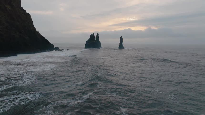 A gray ocean with distant jagged sea stacks at dawn as gentle waves roll toward a rocky shore