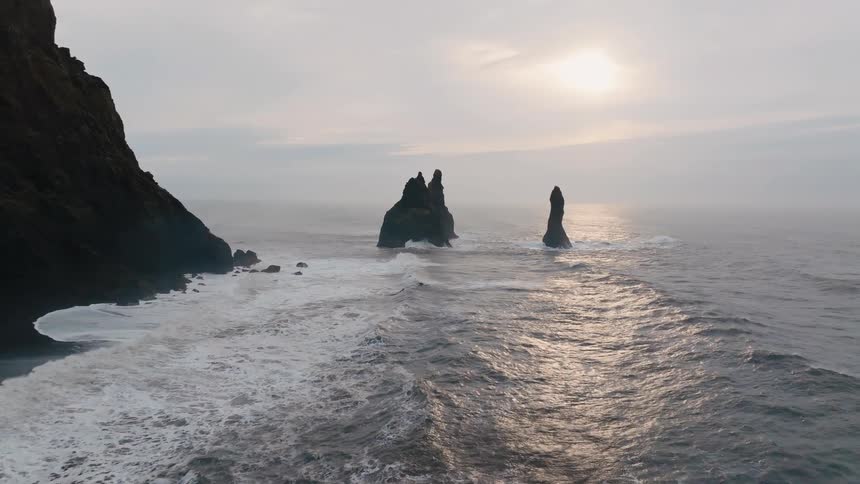 Dramatic basalt sea stacks rise from the choppy ocean as waves crash toward the black cliff shoreline under a bright but hazy sky
