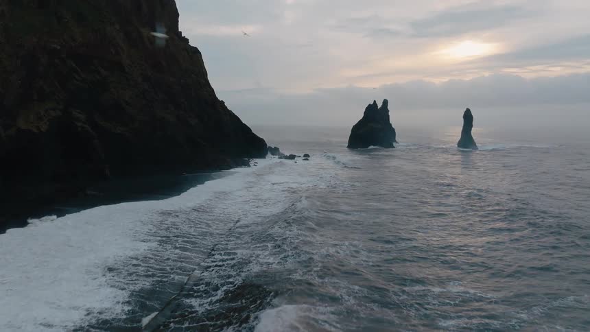Ocean waves crash violently against a rocky shoreline as the golden sun sets in the distance casting warm light across the scene a bird soars overhead silhouetted against the glowing sky