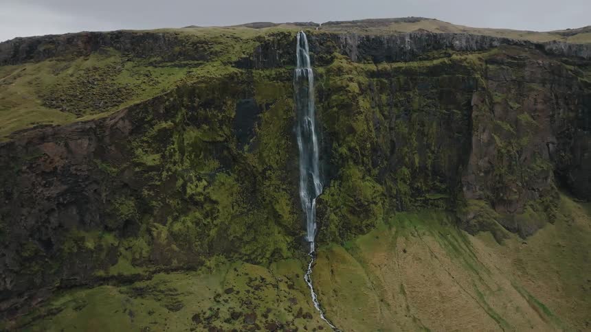 Send a high waterfall plunges over a sheer cliff face covered in bright green moss and grass flowing down to a creek bed below under a cloudy gray sky
