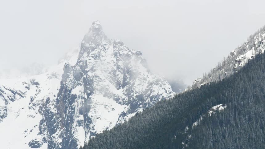 A steep jagged mountain peak covered in snow is visible through fog and partially obscured by a slope covered with dark green forest