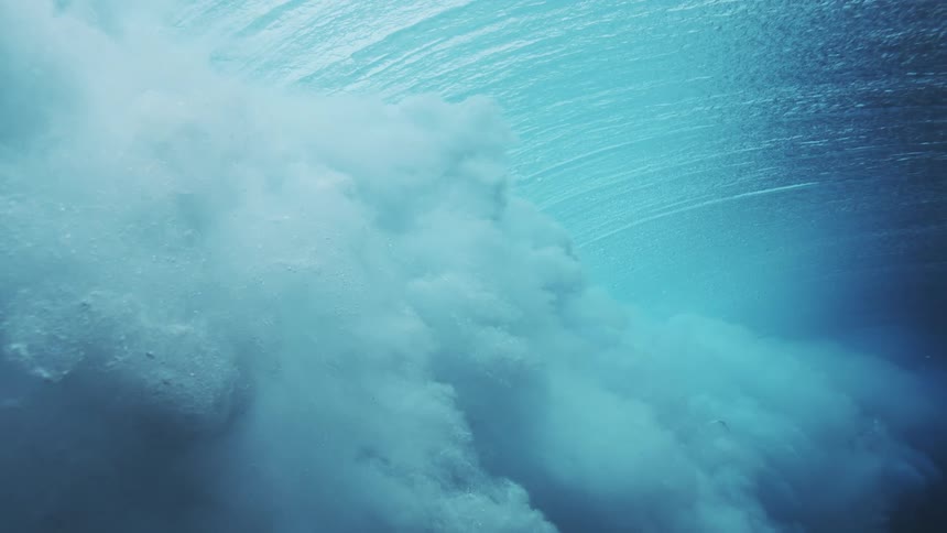 The video is an extreme closeup underwater shot of a large wave breaking and generating a massive amount of bubbles and foam the water is a deep blue and dark turquoise color creating an abstract and powerful depiction of the oceans energy