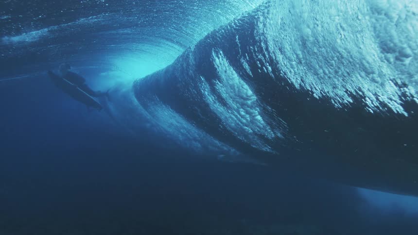 A low underwater view shows a large turquoise ocean wave cresting and breaking with a surfer riding the tube in the distance and natural sunlight filtering through the water