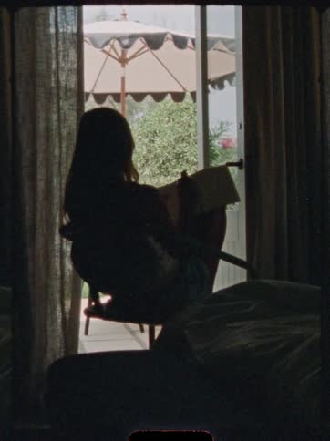 The video is a lowkey indoor shot where a woman is silhouetted against a large window she is seated in a chair by a bed and the bright light reveals an umbrella on a patio outside the window