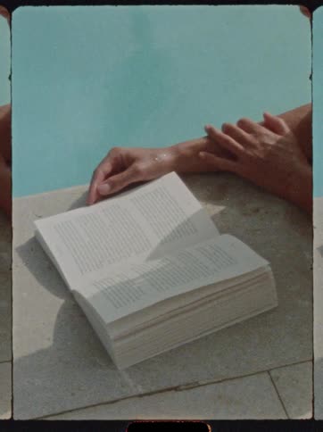 A closeup shot shows an open book resting on the tiled edge of a swimming pool with a persons hands partially visible resting near the book and the blue water