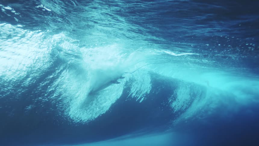 An underwater view captures a surfer amidst the dramatic collapse of a large ocean wave filling the deep blue water with bright light and a massive cloud of foam and bubbles as a surfer duck dives at the bottom