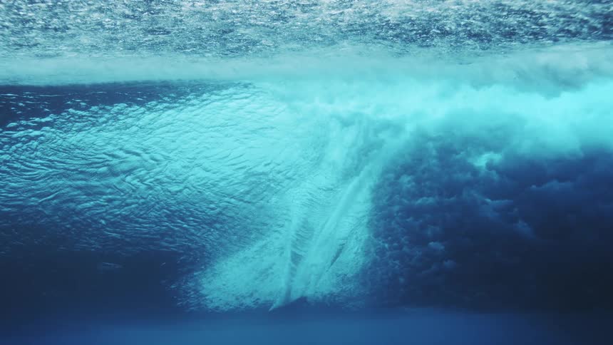 An underwater view captures the base of a surfers board cutting horizontally through a powerful ocean wave that churns into white foam against a deep blue backdrop