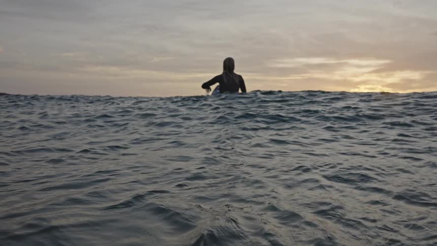 The video shows a person standing in the ocean as the sun sets with the water level reaching their chest the figure is silhouetted against the bright hazy sunset over the water which creates a lowlight tranquil scene