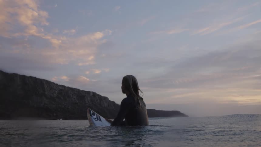 A wide shot shows a woman in a dark wetsuit sitting on a surfboard in the ocean water facing a cloudy sky at sunrise or sunset with a dark cliff and landmass forming a silhouette in the background
