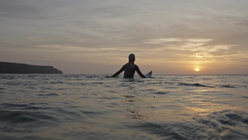 Silhouette of female surfer sitting on surfboard waiting for a wave at dusk