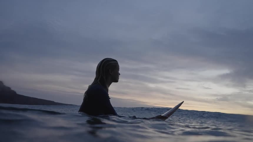 Woman sits on her surfboard watching the sea at dusk