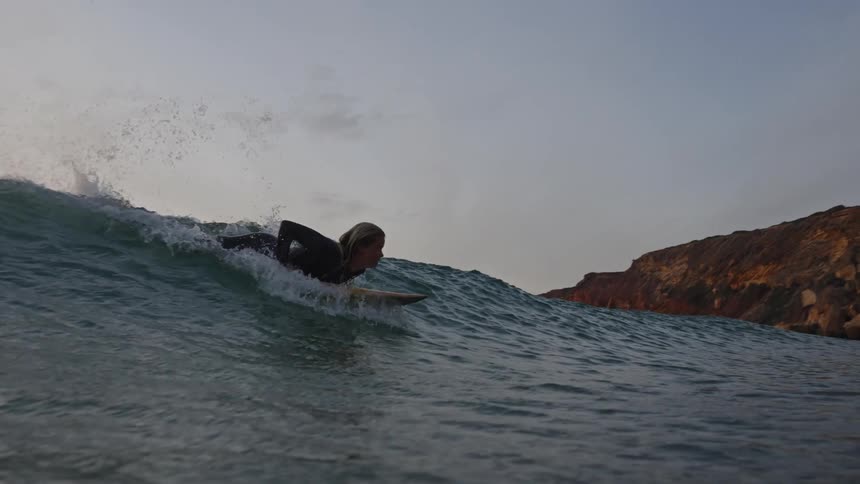 A surfer wearing a dark wetsuit paddles into a breaking ocean wave near a rocky landmass on the right successfully catching the wave and riding its face at sunset before the view goes underwater
