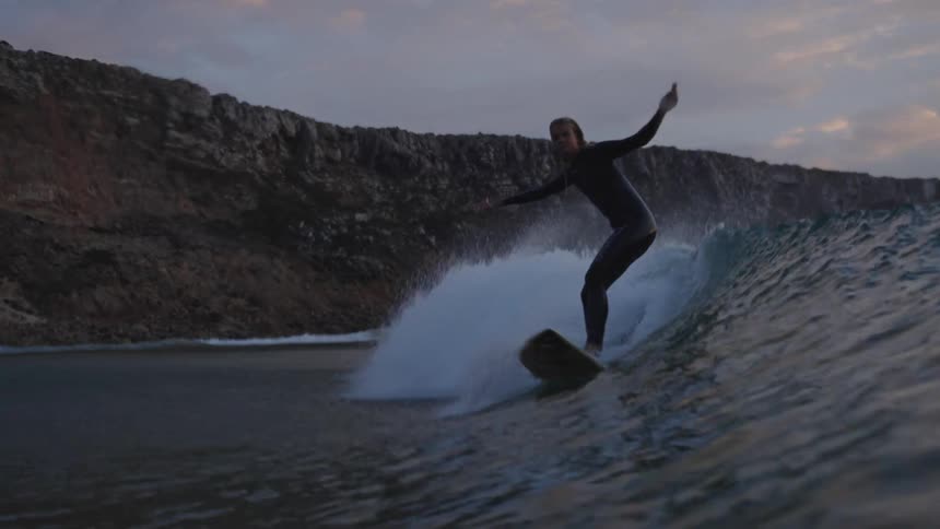 Surfer rides a wave at a beach surrounded by cliffs at sunset
