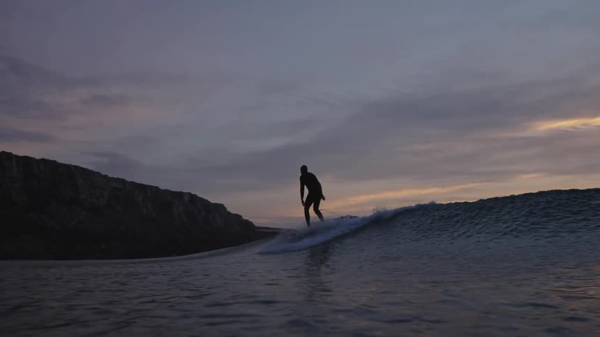 A silhouetted surfer rides a long wave against a dramatic sunset sky