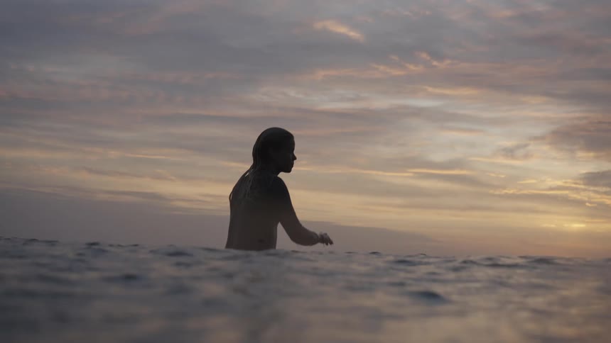 A womans silhouette is visible standing waist deep in the ocean against a pale colorful sunset or sunrise sky as she briefly lifts her hand to touch her wet hair