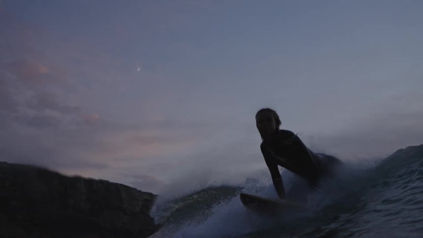 Person surfs in choppy sea during twilight with cliffs in the background under bluishpurple sky