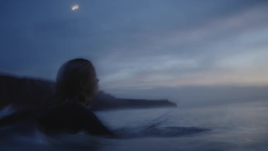 A surfer wearing a dark wetsuit is silhouetted against a darkly overcast dusk sky while paddling on a surfboard into the ocean with the dark outline of a hill or cliff in the background