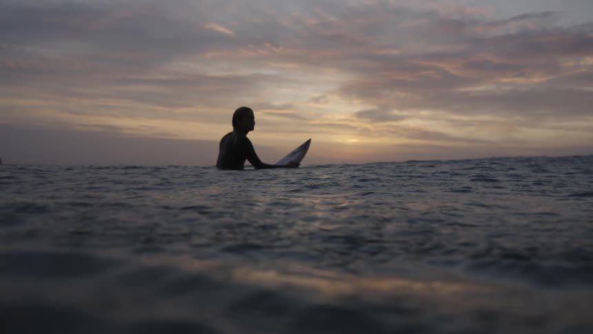 The woman sits on her surfboard at sunrise