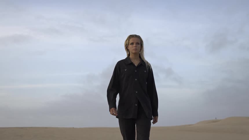 A young woman with long blonde hair walks barefoot across the desert dressed in soft pajamas as the sun casts a warm glow over the sand