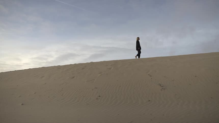 A wide distant view shows the silhouette of a person in dark clothing walking upward along the ridge of a large sand dune against a cloudy pale blue sky