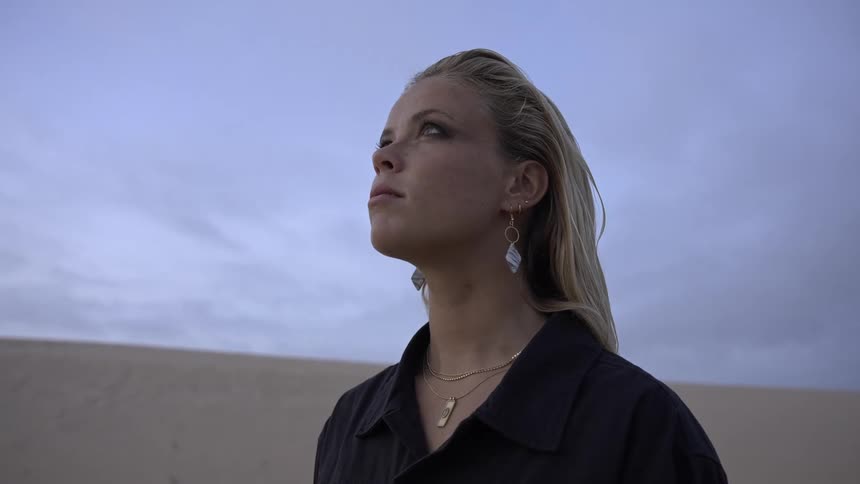 The woman looks up at the cloudy sky while standing on the sand