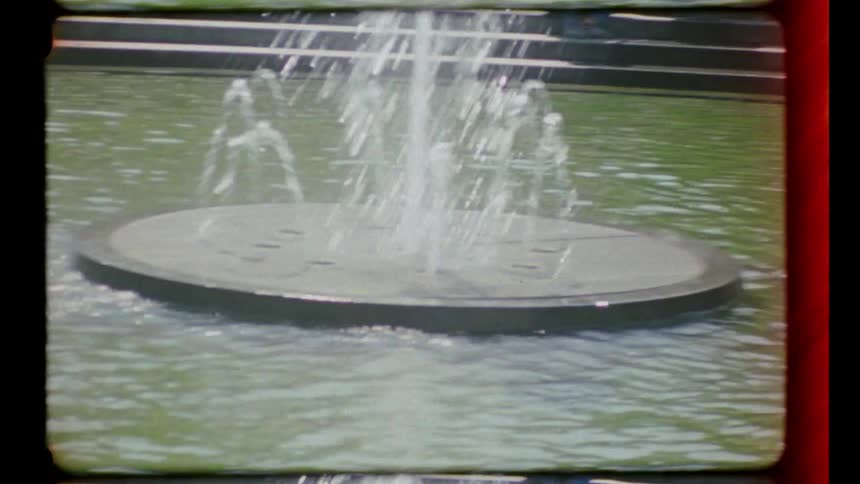 People move around the park near a large fountain on a sunny day