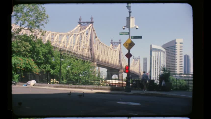 A city skyline at midday with a person walking along a path and a link bridge connecting to another city visible in the distance