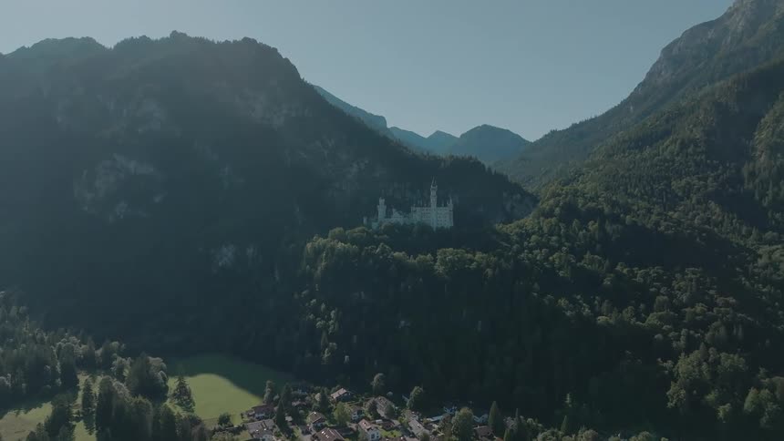 A white hilltop castle sits amid forested mountains above a small village under a clear sky