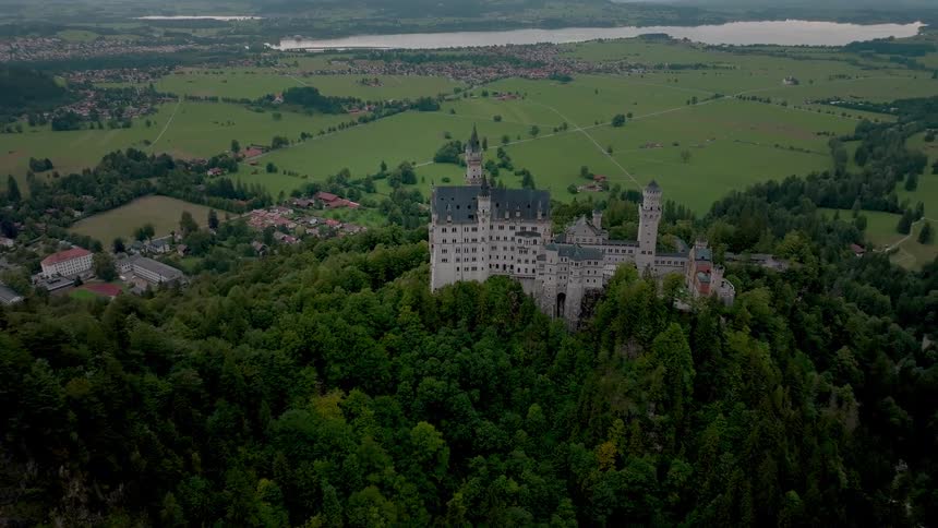 The video is an aerial view revealing the magnificent neuschwanstein castle nestled on a steep wooded hill in bavaria germanythe background shows a panoramic vista of rolling green countryside dotted with villages and large serene lake