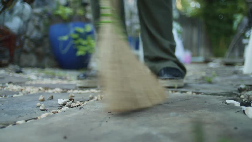 A person sweeps an outdoor stone walkway with a straw broom pushing small pebbles and dirt forward while wearing dark shoes and green pants