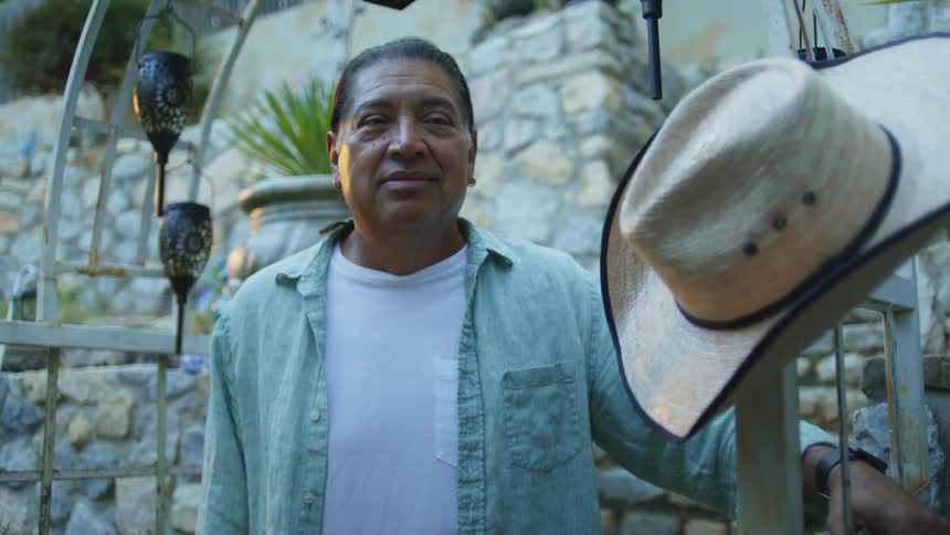A person stands outdoors in front of a stone wall wearing a light blue shirt a straw hat is visible in the foreground hanging lamps and a large planter are visible in the background